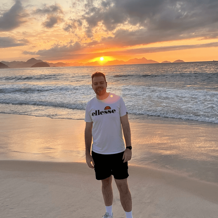 Toby Abram standing on a beach at sunset, in a white T-shirt and black shorts, with the sea behind him.